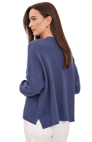 A woman wearing a textured, ribbed knit sweater in a dusty blue shade, shown from a three-quarter back view against a plain off-white studio background.