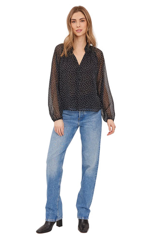 A woman modeling a black long-sleeve blouse with a small white polka dot pattern and sheer balloon sleeves, paired with blue jeans and black ankle boots, against a light gray studio background.