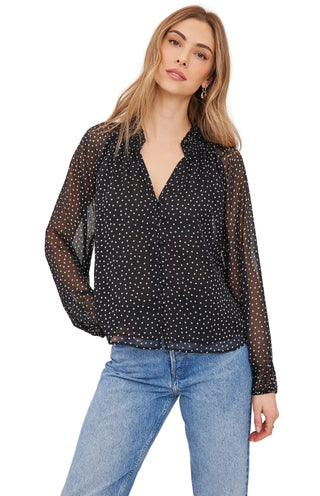 A woman modeling a sheer black long-sleeve blouse with a white polka dot pattern, paired with light blue jeans, against a plain light gray studio background.