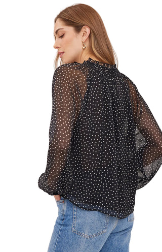 A woman wearing a sheer black long-sleeve blouse with white polka dots, shown from a side-back angle against a plain light background.