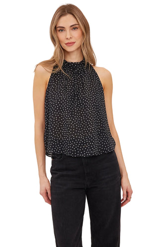 A woman modeling a sleeveless black high-neck top with a white polka dot pattern, paired with black pants, against a plain light gray studio background.