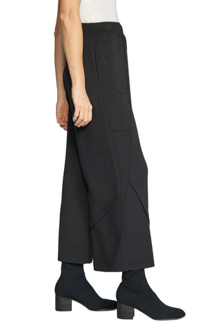 A model is shown from the waist down wearing black wide-leg cropped trousers with diagonal seam details, paired with black ankle boots against a light gray studio background.