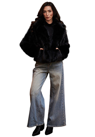Woman wearing a black fur coat and blue jeans standing in front of a red sofa with a painting in the background.