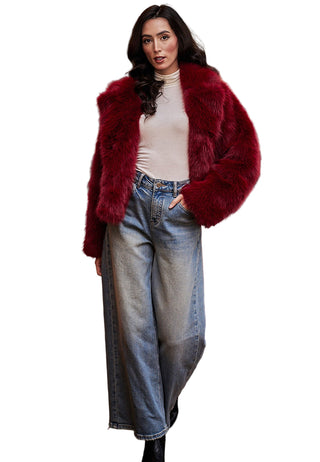 Woman wearing a red fur coat, white top, and blue jeans standing in front of a red velvet couch.
