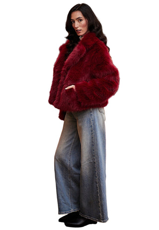 Woman wearing a red fur coat and blue jeans standing in front of a red sofa and painting.