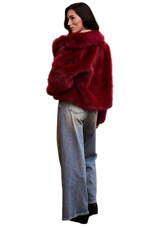Person wearing a red fur coat standing in front of a red sofa with a painting in the background.