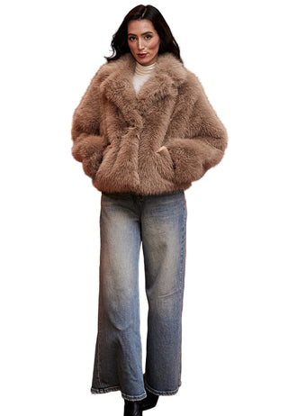 Woman wearing a brown fur coat and jeans standing in front of a red sofa with a painting in the background.