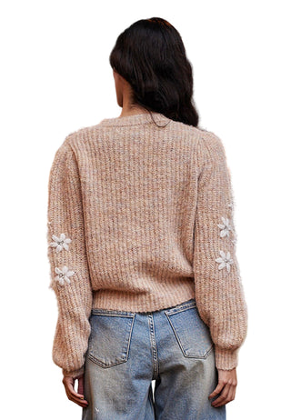 Person wearing a beige knitted sweater with floral embellishments, standing in front of a decorative wall.