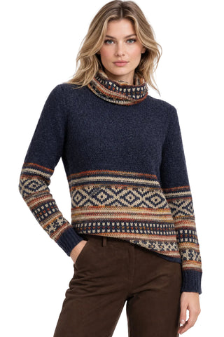 A woman wearing a navy blue knit turtleneck sweater with a multicolor geometric Fair Isle pattern across the chest, cuffs, and lower hem, paired with brown pants against a white background.