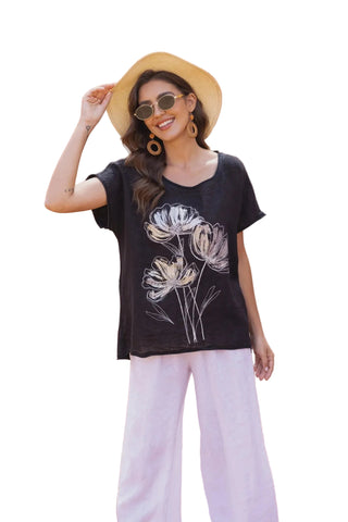 Woman wearing a black t-shirt with floral design and white pants, standing in front of a textured wall.