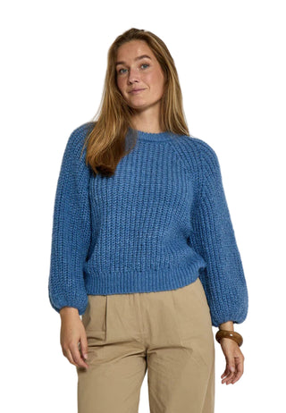 Woman wearing a blue knitted sweater and beige pants against a white background