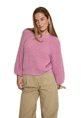 Woman wearing a pink knitted sweater and beige pants sitting on a wooden chair against a white background