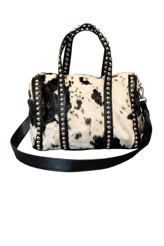 A cow-print handbag with black faux-leather trim and gold-tone studs, sitting on a gray countertop in an indoor setting.