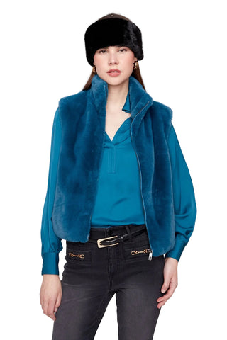 A model wearing a teal blue faux fur vest over a matching teal top, paired with a black fur hat and dark gray jeans, against a light gray studio background.