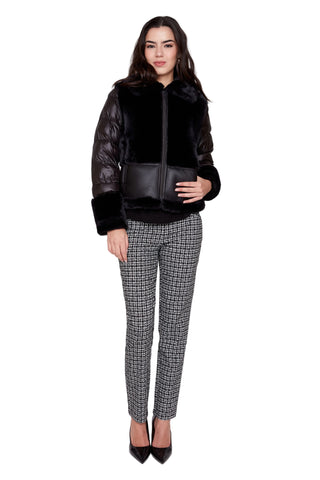 A model stands against a light gray studio background wearing a black patchwork jacket with faux fur trim, black leather sleeves, and a black zippered front. She is also wearing black and white houndstooth patterned trousers and black pointed-toe heels.