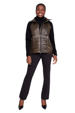 A woman modeling a leopard-print faux-leather puffer vest over a black turtleneck and black pants, standing against a light gray studio background.