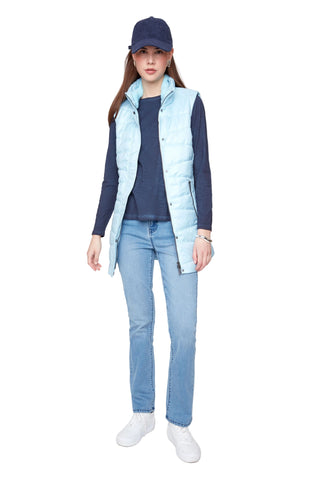 A woman modeling a light blue puffer vest over a navy long-sleeve top, paired with light blue jeans, white sneakers, and a navy baseball cap, against a plain light gray studio background.