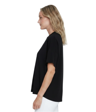 A woman in profile wearing a loose black short-sleeve top and dark leggings, standing against a plain light gray studio background.