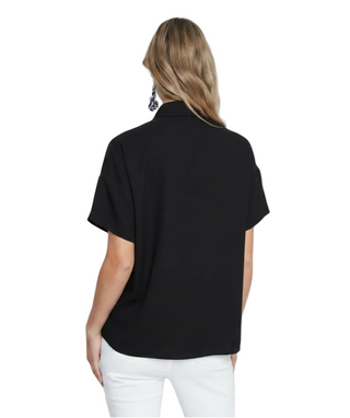 A woman viewed from the back wearing a black short-sleeve collared top and dark pants against a plain light gray studio background.