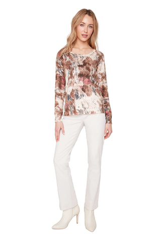 A woman modeling a long-sleeve crewneck top with an abstract brown, beige, and pink pattern, paired with off-white trousers and matching ankle boots, against a light gray studio background.