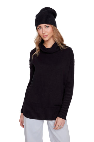 A woman wearing a black ribbed knit cowl-neck sweater, a black beanie, and white pants, photographed against a light gray studio background.