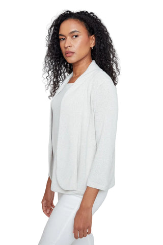 A woman with curly dark hair wearing a light off-white textured cardigan over a white top and white pants, photographed against a plain light gray studio background.