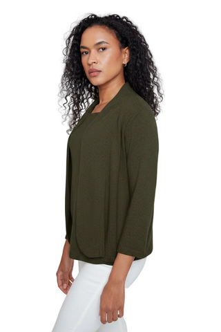 A woman with curly black hair wearing an olive green long-sleeve top and white pants, photographed against a light gray studio background.