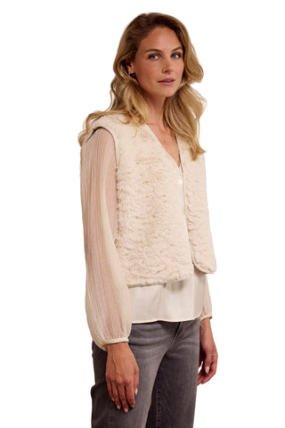 Woman wearing a cream lace vest over a white blouse with gray pants on a beige background