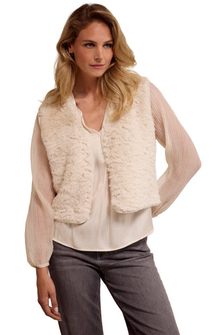 Woman wearing a cream-colored vest over a white blouse and gray pants on a plain background