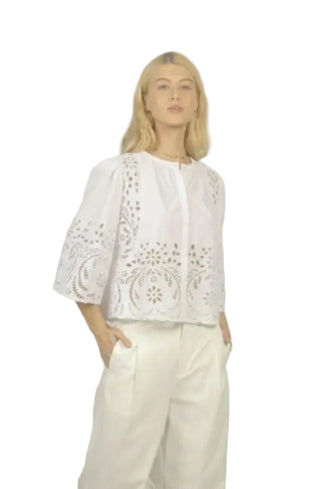 Woman wearing a white blouse with intricate patterns and white pants against a plain background