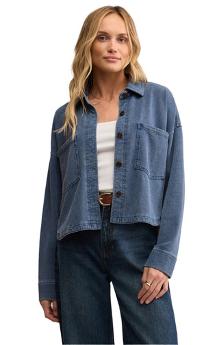 Woman wearing a blue denim jacket over a white top and dark jeans on a beige background