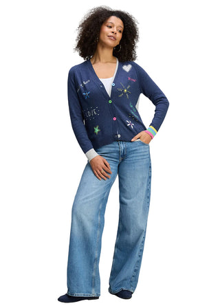 Woman wearing a blue cardigan with colorful designs and blue jeans on a gray background