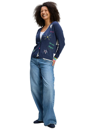 Woman wearing a blue cardigan with colorful designs and text, paired with blue jeans on a gray background