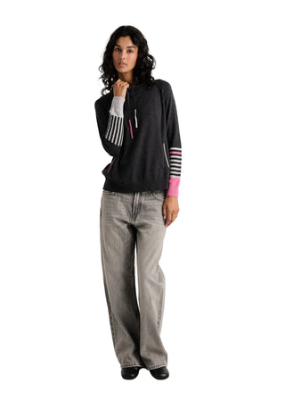 A woman modeling a dark gray long-sleeve knit top with striped cuffs in black, white, and pink, paired with light gray wide-leg jeans and black shoes, against a plain light gray studio background.