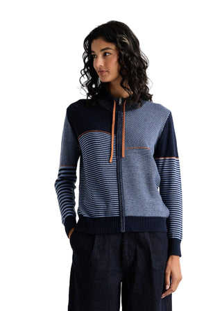 A woman modeling a navy and blue striped zip-up sweater with textured panels and orange zipper pulls, paired with dark trousers, against a plain light gray studio background.