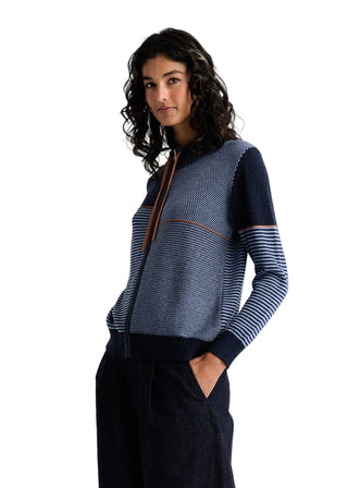 A woman modeling a color-blocked knit zip-up cardigan with navy, blue, and white stripes, paired with dark navy trousers, against a plain light gray studio background.