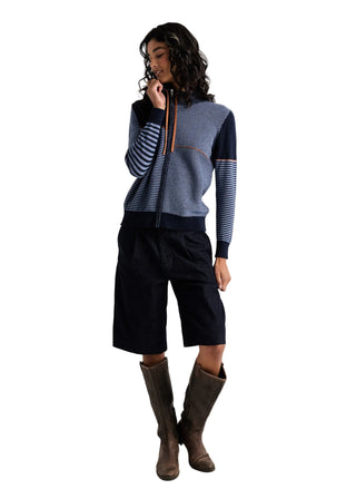 A model wearing a color-blocked knit zip-up sweater with navy, slate blue, and striped sleeves, paired with dark navy cropped pants and brown knee-high boots, against a light gray studio background.