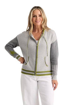 A woman modeling a light gray and heather gray colorblock zip-up hoodie with olive green accents, including drawstrings, pocket trim, and ribbed hem and cuffs, paired with white pants against a light gray studio background.