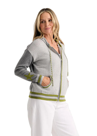 A woman modeling a light gray zip-up hoodie with contrasting dark gray sleeves and green striped trim, paired with white pants, against a plain light gray studio background.