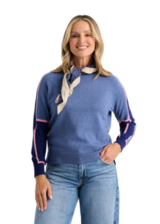 A woman modeling a blue knit sweater with navy sleeves featuring pink stripes, styled with a cream and navy patterned neck scarf and blue jeans against a light gray studio background.