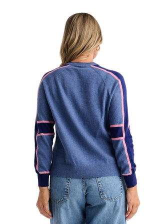 A woman viewed from the back wearing a blue knit sweater with pink contrast trim and navy side panels, paired with light blue jeans, against a plain light gray studio background.
