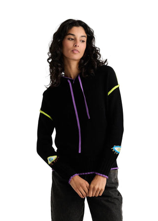 A woman wearing a black knit pullover hoodie with purple drawstrings, neon yellow-green sleeve stripes, and colorful embroidered patches on the cuffs and chest, paired with dark gray jeans against a light gray studio background.
