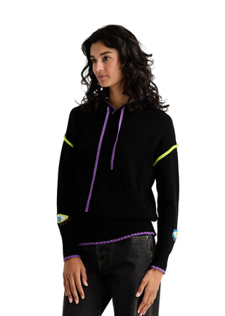 A woman modeling a black knit hoodie with purple drawstrings, neon yellow-green sleeve stripes, and purple trim details, paired with dark jeans against a light gray studio background.