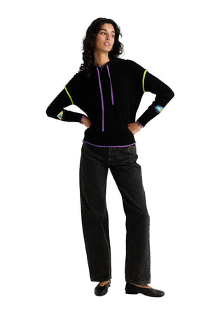 A woman modeling a black knit long-sleeve top with neon green and purple accents, paired with black jeans and black flats, against a light gray studio background.