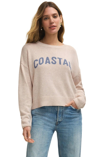 Woman wearing a beige sweater with 'COASTAL' text and blue jeans on a plain background
