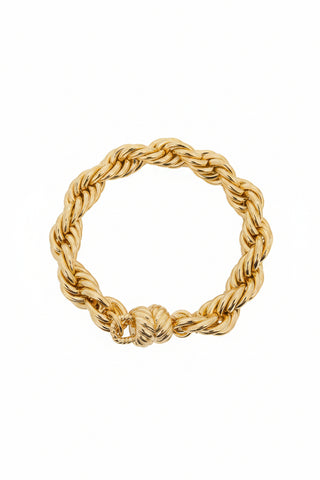 A gold rope-style chain bracelet displayed on a black jewelry stand, with a textured light gray background featuring circular reflections.