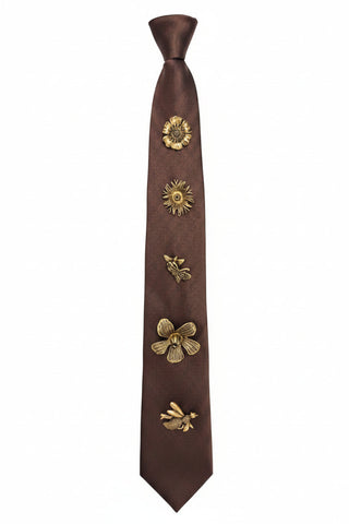 A dark brown satin necktie displayed on a black mannequin torso, decorated with five metallic gold insect and floral appliqués running down the center. The background features a textured metallic silver circular pattern.