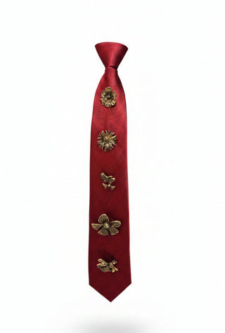 A red necktie displayed on a black mannequin torso, decorated with five metallic gold floral and insect-shaped pins arranged vertically down the front.