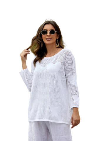 Woman wearing a white outfit with sunglasses and earrings, standing outdoors with greenery in the background.