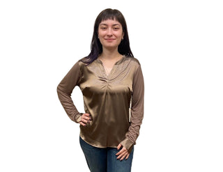 Woman wearing a brown long-sleeve top and blue jeans standing against a beige curtain.